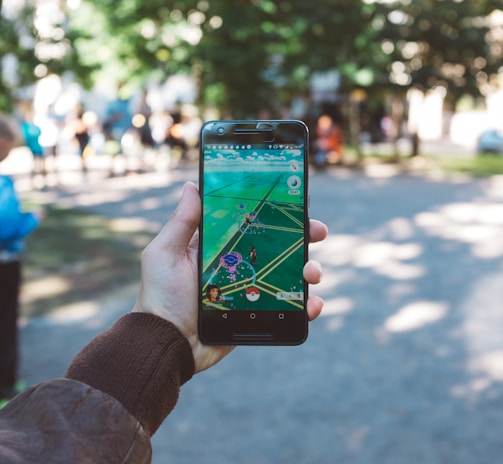 A person is holding a smartphone with a gaming app open, featuring an augmented reality map. The setting is outdoors, with people visible in the background, suggesting a park or public space.